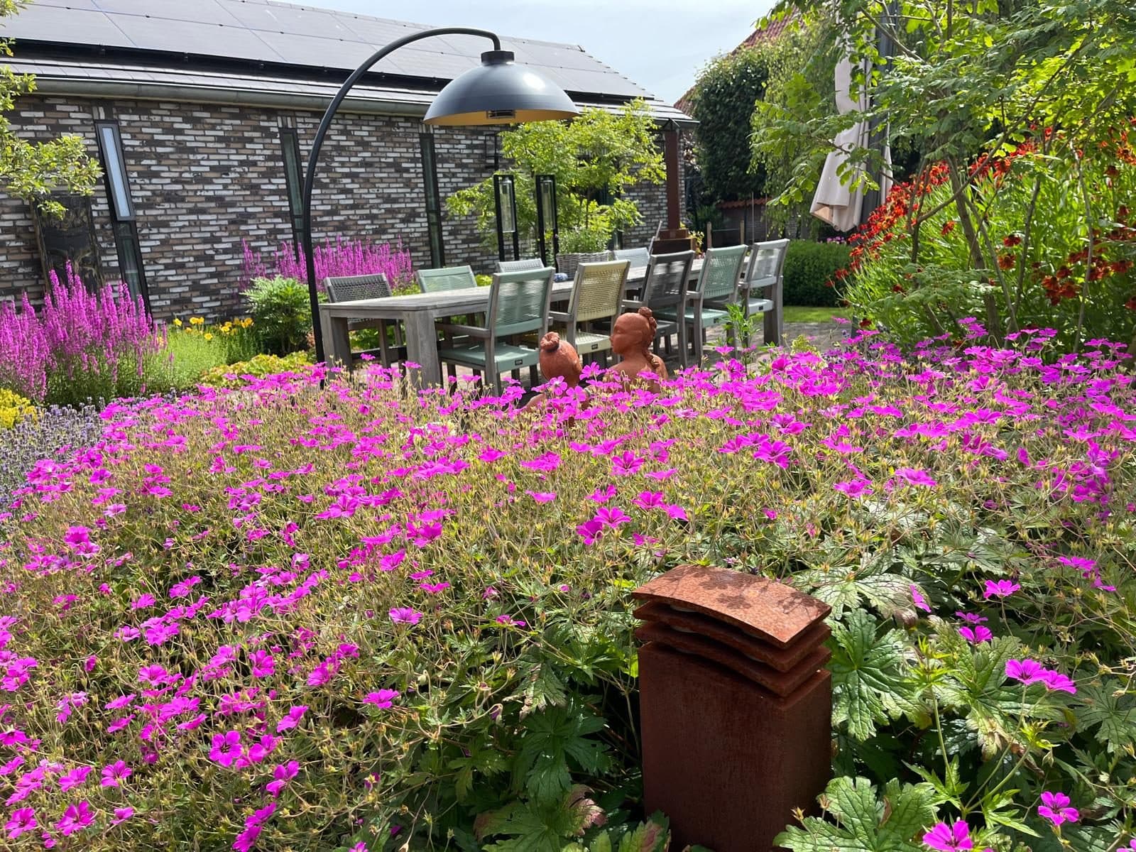 Een terras van gebakken klinkers met borders vol roze geraniums ernaast