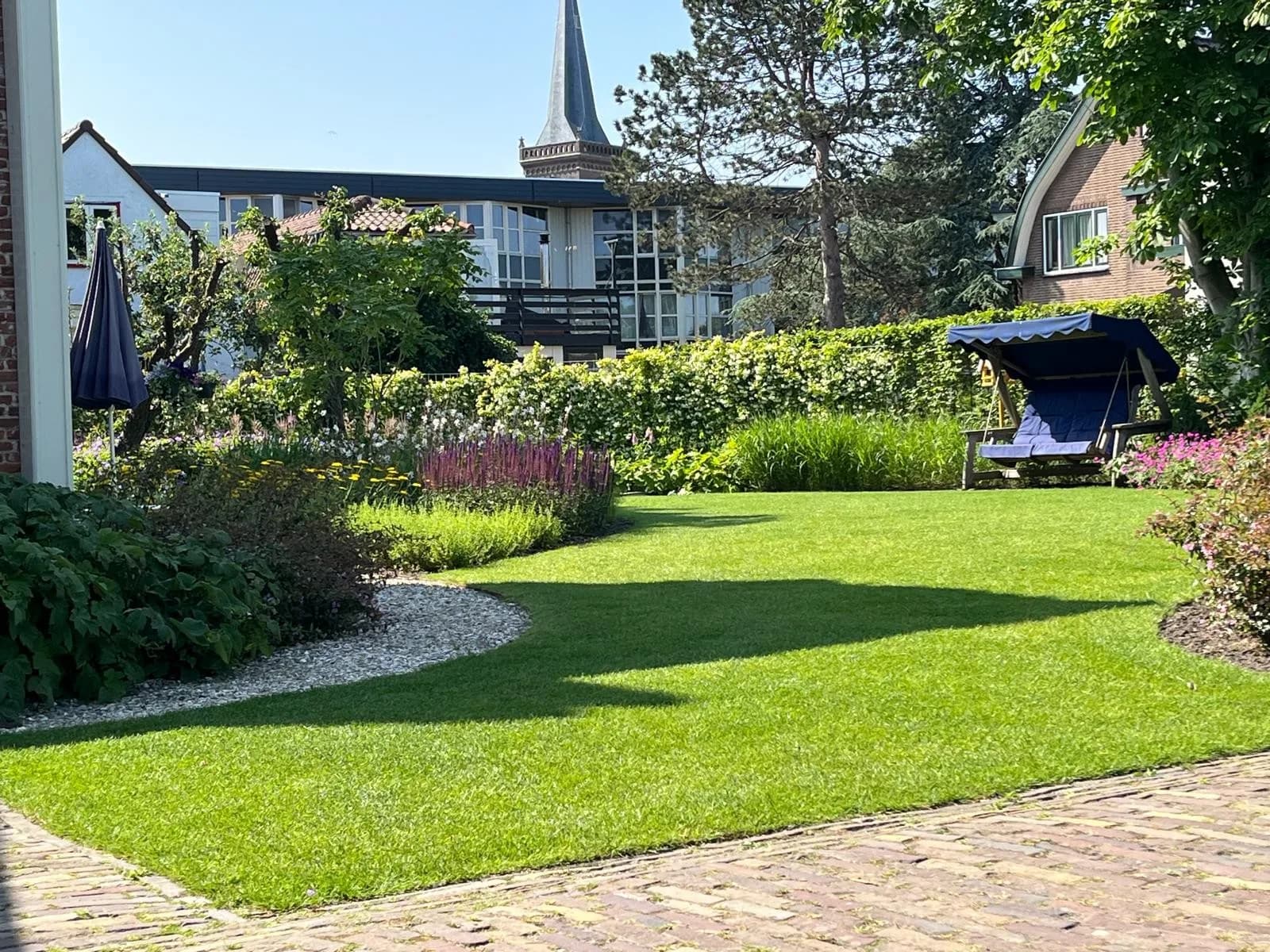 Gazon met borders en een schommelbank, met een kerktoren op de achtergrond
