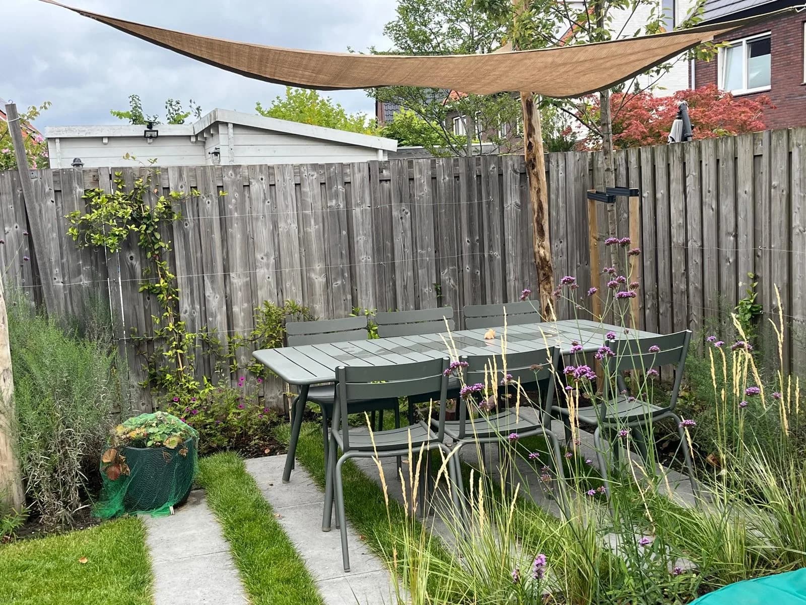 Terras onder een schaduwdoek, met siergrassen en betonplaten in het gazon